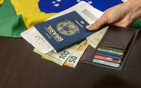 Mão segurando passaporte brasileiro sobre mesa com bandeira brasileira ao fundo