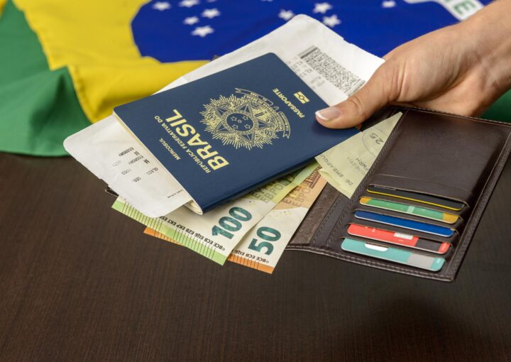 Mão segurando passaporte brasileiro sobre mesa com bandeira brasileira ao fundo