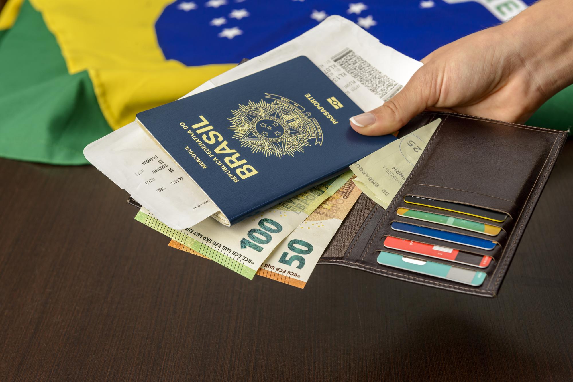 Mão segurando passaporte brasileiro sobre mesa com bandeira brasileira ao fundo